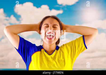 Giovane donna ecuadoriana indossando maratona ufficiale Football Shirt in piedi di fronte alla fotocamera, molto impegnato il linguaggio del corpo la visione di gioco con grande entusiasmo, cielo blu e nuvole sullo sfondo Foto Stock