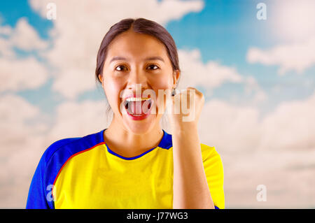 Giovane donna ecuadoriana indossando maratona ufficiale Football Shirt in piedi di fronte alla fotocamera, molto impegnato il linguaggio del corpo la visione di gioco con grande entusiasmo, cielo blu e nuvole sullo sfondo Foto Stock