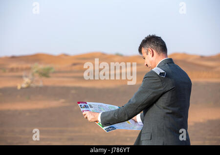 Imprenditore con una valigia in un deserto, guardando una mappa per raggiungerci Foto Stock