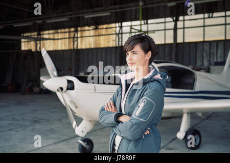 Fiducioso giovane pilota femmina in posa di un hangar con le braccia incrociate, aereo sullo sfondo Foto Stock