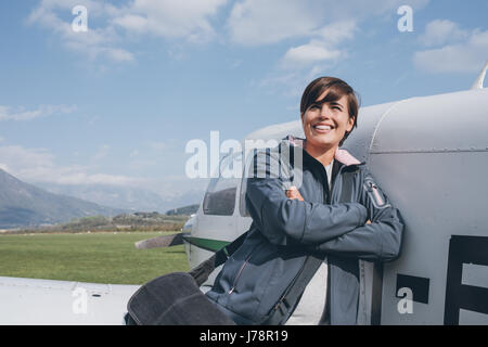 Sorridendo allegro pilota femmina appoggiata su un aeroplano ad elica e guardando lontano, cielo blu sullo sfondo, viaggi e concetto di aviazione Foto Stock