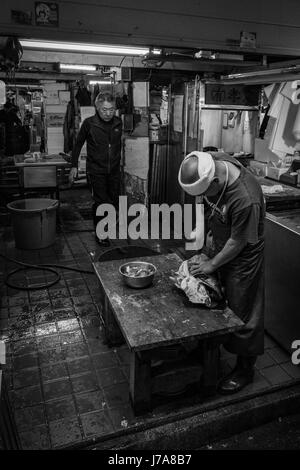 Foto in bianco e nero di due uomini che lavorano nel loro box presso il mercato del pesce Tsukiji. Il pescivendolo è lo svuotamento e la pulizia di una grande testa di pesce. Foto Stock