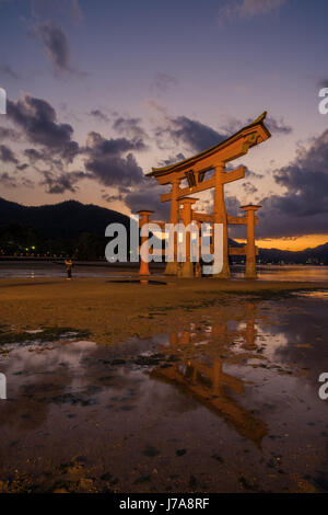 Si tratta di bassa marea e un uomo è tenuto un selfie dal Miajima torii. Il cielo è viola e golden e il torii è riflessa in una pozza d'acqua. Foto Stock