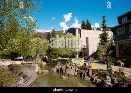 Il Whistler in una giornata di sole in primavera. Persone sedersi dal villaggio di Whistler corso d'acqua che scorre accanto al Maury giovani Arts Centre Whistler BC, Foto Stock