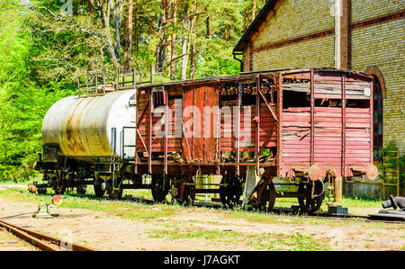 Abbandonato red vintage boxcar di legno in piedi sul binario ferroviario all'esterno in mattoni gialli con auto chimica e le foreste in background. Foto Stock