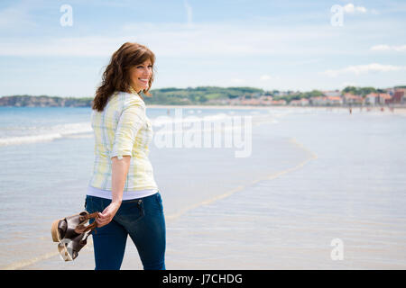 Attraente e felice donna di mezza età camminando lungo la riva del mare tenendo le scarpe in mano Foto Stock
