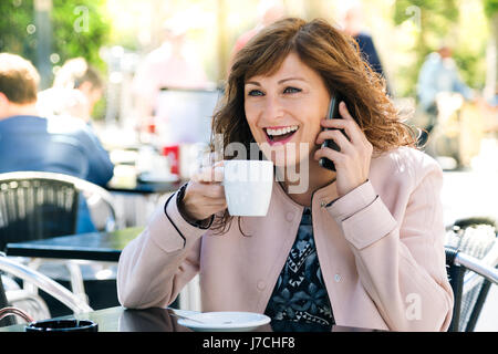 Bella e felice donna di mezza età ridere del telefono cellulare mentre un caffè all'aperto Foto Stock