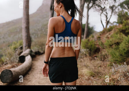 Vista posteriore del colpo di felice giovane donna asiatica camminando sul sentiero di montagna e sorridente. Donna Fitness in sportswear su cross country percorso. Foto Stock