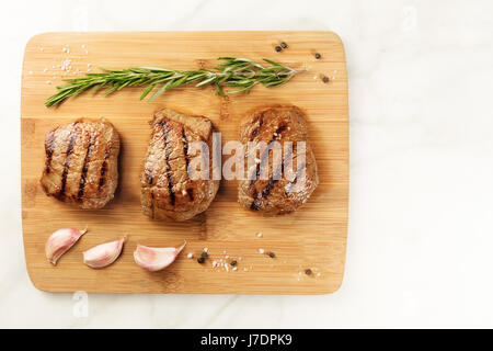 Tre fette di carne alla brace, filetti di manzo, ripresa dall'alto su una tavola di legno con un rametto di rosmarino, 4 spicchi di aglio, sale e pepe Foto Stock