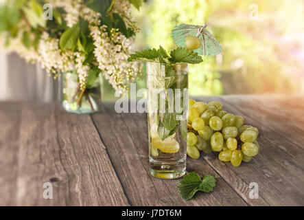 Detox acqua menta, succo d'uva e il ghiaccio in un vetro su uno sfondo di un mazzo di fiori di ciliegio e uva in una giornata di sole. Il telaio orizzontale. Foto Stock