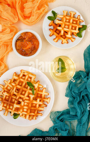 Waffle belga su piastre guarnite con foglie di menta, coppa di vetro con il tè verde con menta e confettura di pesche con il rosmarino in una piccola ciotola su sfondo beige Foto Stock