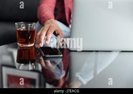 Immagine ritagliata del giovane uomo seduto a casa all'interno mentre si utilizza il computer portatile tenendo il telefono. Foto Stock