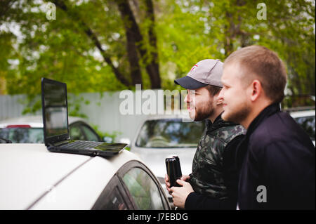 Uomo con notebook nel parcheggio nel cortile vicino a macchina sta facendo manipolazioni con cyber sistema, concetto Foto Stock