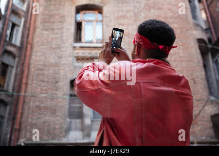 Vista posteriore ritratto di una giovane ragazzo africano nella fascia di scattare una foto con il telefono cellulare all'aperto Foto Stock