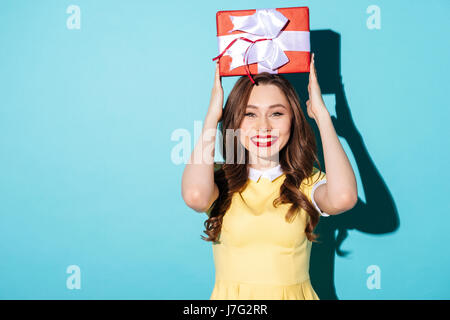 Ritratto di una bruna sorridente ragazza in abito tenendo presente la casella sopra la sua testa e guardando la telecamera isolate su sfondo blu Foto Stock