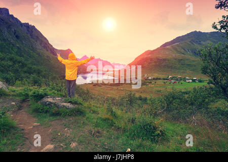Vista panoramica del fiordo. Twilight time con un incredibile cielo arancione. Il giovane uomo con le mani in aria in piedi su una rupe di roccia e guardando suns Foto Stock