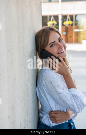 Femmina e giovane studente universitario sul campus universitario con il suo smartphone e sorridente in piedi fuori vicino a muro con spazio di copia Foto Stock