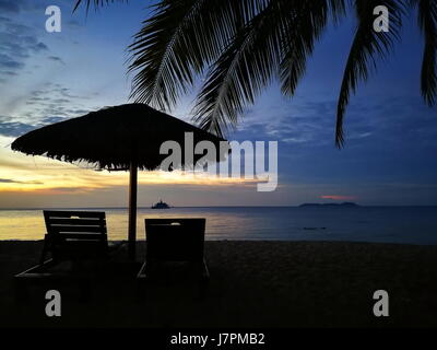 Tramonto al mare in un'isola tropicale, orlate da alberi di cocco ondeggianti nella brezza del mare e con silhouette di gazebo e sedie a sdraio. Foto Stock