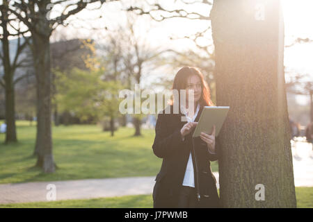 Business donna in nero business suit holding tablet pc in piedi nel parco contro la luce del sole Foto Stock