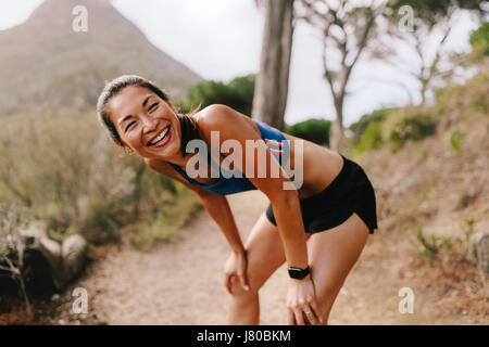 Sano giovane donna in piedi sul sentiero di montagna con le sue mani sulle ginocchia e ridere. Asian femminile in sportswear prendendo una pausa dopo aver eseguito wo Foto Stock