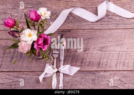 Inserire le impostazioni di una tavola con coltello e forchetta, legata con un bianco di raso e fiori su un tavolo vintage vista superiore, piana, laici vista aerea Foto Stock