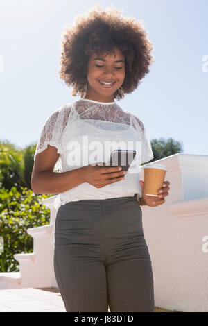 Donna sorridente con il telefono cellulare mentre si tiene tazza monouso contro sky sulla giornata di sole Foto Stock