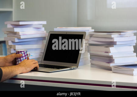 L'uomo che lavora su un computer portatile con uno schermo vuoto in biblioteca con le pile di libri intorno tenendo una tazza da caffè Foto Stock