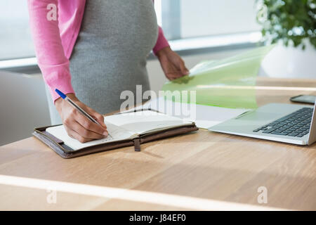 La sezione centrale della gravidanza imprenditrice iscritto in agenda elettronica alla scrivania in ufficio Foto Stock