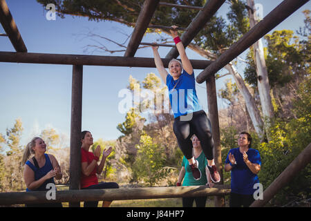 Donna di essere acclamato bye i suoi compagni di squadra a salire Monkey bar durante la corsa a ostacoli della formazione in boot camp Foto Stock