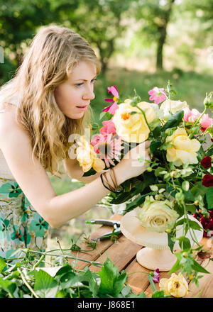 Celebrazione, Wedding, flora, la bellezza del concetto di natura - bionda blue-eyed giovane donna organizza composizione floreale in supporto di legno con aiuto del giardino potatore. bouquet contiene inglese rose e peonie. Foto Stock