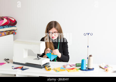 Ricamo e la trapuntatura in officina di una giovane donna - una donna su misura ad una scrivania e della macchina per cucire di maglie con mano i lembi di tessuto colorato che giace accanto al thread, tessuti, aghi, spilli. Foto Stock