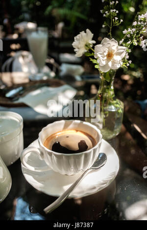 Pasto all'aperto nel giardino sotto la luce del sole passante da alberi con una tazza di caffè nero collocato sul tavolo di vetro accanto a un barattolo di vetro riempito con stagno Foto Stock