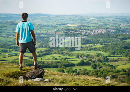 Uomo in pantaloncini e maglietta in piedi sulla roccia di piccole dimensioni che si affaccia sulla valle. Brecon città e campi in background. Girato in Galles nella tarda primavera. Foto Stock