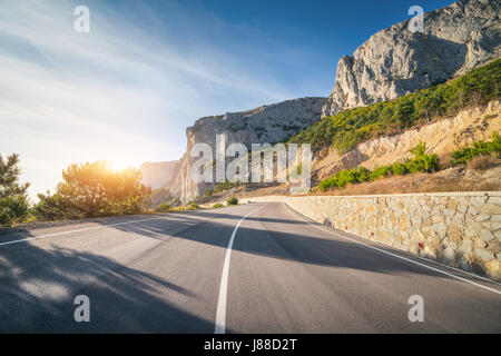 Strada asfaltata in estate a sunrise. Paesaggio con vuoto bella strada di montagna con un asfalto perfetto, alte rocce, alberi e sunny blue sky. Travel b Foto Stock