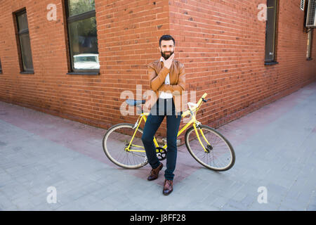 Giovane uomo barbuto tenendo la mano sul mento mentre è seduto vicino la sua bicicletta all'aperto vicino a un muro di mattoni Foto Stock