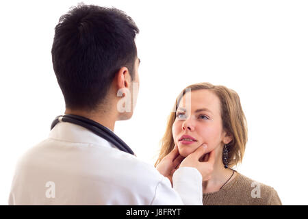 Giovane medico con i capelli scuri in abito bianco con uno stetoscopio sul suo collo esaminato piacevole giovane donna con capelli lunghi marrone in maglione e jeans su bianco Foto Stock