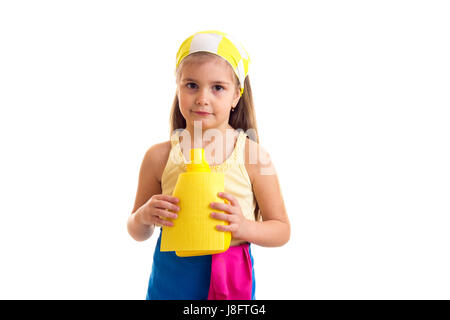 Graziosa bambina con lunghi capelli castani in maglietta gialla e grembiule blu con rosa guanti e fazzoletto giallo giallo holding duster e bottiglia di colore giallo Foto Stock