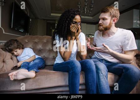 Nero donna piangere mentre suo marito parla con lei. Famiglia comodamente seduto sul divano. Coppia con il loro piccolo figlio. Foto Stock