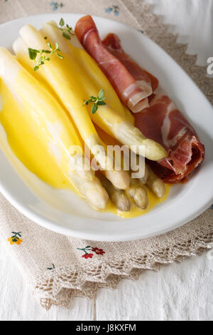 Ricca prima colazione: asparagi bianchi serviti con salsa olandese e prosciutto close-up su una piastra sul piano verticale. Foto Stock