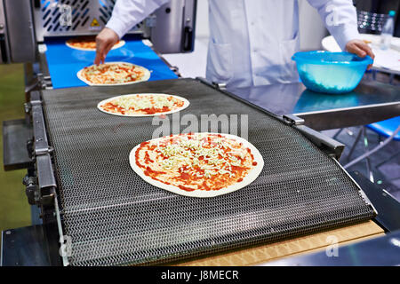 Lavoratore baker cucinare aggiunge il Formaggio per pizza sul trasportatore Foto Stock