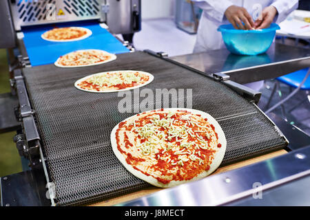 Lavoratore baker cucinare aggiunge il Formaggio per pizza sul trasportatore Foto Stock