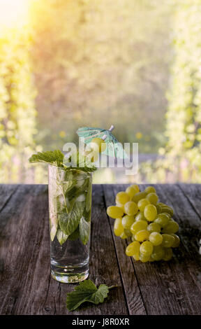 Detox acqua menta, succo d'uva e il ghiaccio in un bicchiere su sfondo verde bokeh e uva in una giornata di sole. Il telaio verticale. Foto Stock