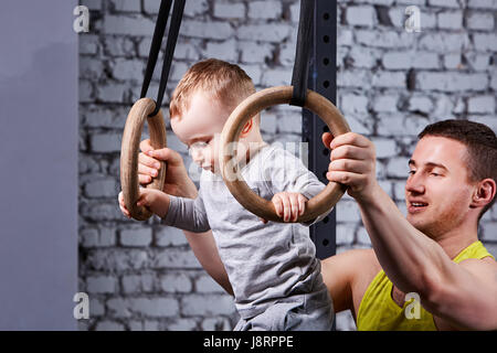 Ritratto di felice giovane padre treni il piccolo figlio con anelli gimnastic contro un muro di mattoni in palestra. Foto Stock