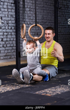 Sorridente giovane padre treni il piccolo figlio con anelli gimnastic contro un muro di mattoni in palestra. Foto Stock