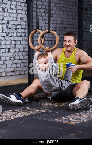 Sorridente giovane padre treni il piccolo figlio con anelli gimnastic contro un muro di mattoni in palestra. Foto Stock