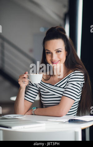 Ritratto di una donna affascinante seduti a tavola e tenendo una tazza di tè guardando la telecamera. Foto Stock