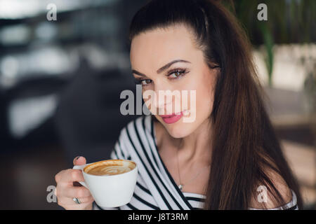 Close-up ritratto di una donna affascinante tenendo una tazza di cappuccino e guardando la telecamera. Foto Stock
