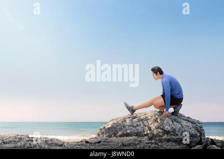 Ritratto di asiatici uomo fitness gambe stretching prima di fare jogging sulla spiaggia Foto Stock