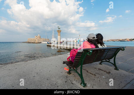RHODES, Grecia - 13 giugno 2015: la gente seduta su una panchina di Mandrachi porto cervo osservando statue e Forte di San Nicola con il faro Foto Stock