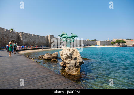 Rodi, Grecia- Agosto 20, 2015; Peopla a piedi nelle vicinanze della statua di delfini a Kolona al di fuori del porto di Rodi città vecchia. Rhodes, Grecia Foto Stock